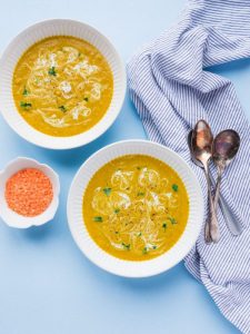 two bowls of curry turmeric lentil soup with a side of lentils next to spoons and napkin in a blue backdrop.